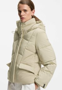 Beige puffjakke med hætte, der har chevron-quilting, to forlommer og elastiske manchetter for ekstra varme og komfort.