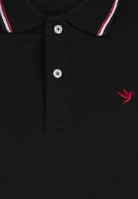 Camisa polo negra con tejido texturizado, tapeta de tres botones y un logo de pájaro bordado en rojo. El cuello tiene rayas en rojo y blanco.