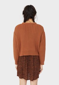 Cropped ribgebreide trui in gebrande oranje, gecombineerd met een doorschijnende zwarte jurk met bruin luipaardprint. Lange, losse mouwen met brede manchetten.