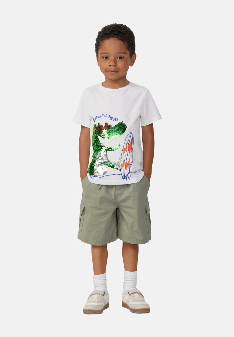 Weißes Baumwoll-T-Shirt mit einem Grafiken eines Alligators aus grünen und silbernen Pailletten, einem orangefarbenen Surfbrett und dem Text "CATCH THE WAVE!", kombiniert mit khakifarbenen Shorts.