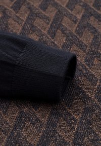 Schwarzer gestrickter Stoffärmel, der sich über einen strukturierten braunen Hintergrund mit einem geometrischen Muster erstreckt. Der Ärmel ist gerippt und hat eine glatte Oberfläche.