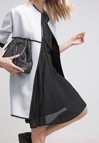 Světloměděný kabát s černým lemem, přes černé skládací šaty. Černá taška s šňůrkou a cvočky. Hladké textury, elegantní silueta.