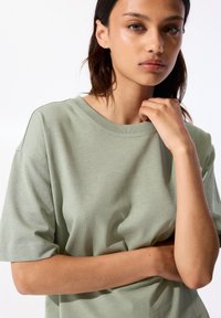T-shirt en coton vert clair avec un col rond et des manches courtes. Le tissu semble doux avec une texture subtile et une coupe décontractée.