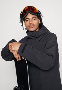 Schwarze isolierte Snowboardjacke mit hohem Kragen und verstellbaren Bündchen. Die Person hält ein Snowboard und trägt orangetönte Skibrille.