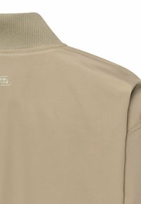 Beżowa kurtka z prążkowanym kołnierzem i małym białym logo na górnej części pleców, pokazująca gładką tkaninę i detale szwów na ramieniu.