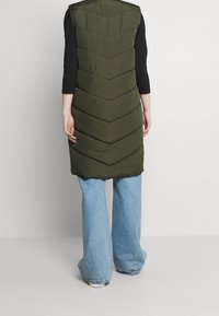 Długa oliwkowo-zielona pikowana kamizelka z przeszyciami w kształcie jodełki. Stylizowana z czarną bluzką z długim rękawem i jasnoniebieskimi dzwonowatymi jeansami.