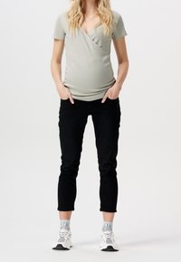 Světle šedý těhotenský top s překládaným designem, krátkými rukávy a texturovanou látkou, kombinovaný s černými skinny džíny a bílými sportovními botami.