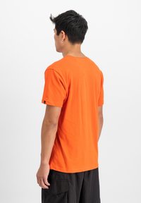 Camiseta de algodón naranja de manga corta con escote redondo, con una etiqueta en la manga izquierda y un ajuste relajado, mostrada desde la parte de atrás.
