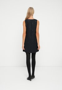 Femme avec de longs cheveux blonds se tenant de dos, portant une robe noire texturée sans manches, des collants noirs et des talons noirs.