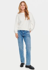 Maglione bianco, con texture, collo rotondo e delicate decorazioni sul davanti, abbinato a jeans dritti blu chiaro e scarpe nere.