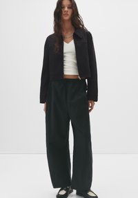 Jachetă scurtă neagră cu guler înalt, cămașă albă cu decolteu în V dedesubt, pantaloni verde largi și pantofi negri cu un design texturat.