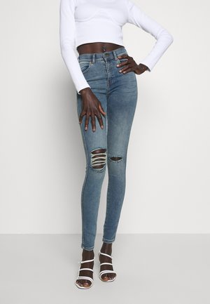 Personne à la peau foncée portant un jean skinny bleu déchiré, un haut blanc à manches longues court et des talons ouverts à brides blancs, debout devant un fond blanc.