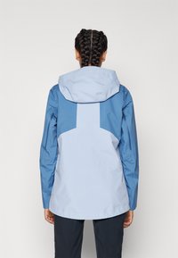Chaqueta impermeable azul con capucha de un azul más claro, mangas raglán y un diseño distintivo en el panel trasero. Textura suave con acentos mínimos.