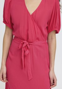 Femme portant une robe portefeuille rose texturée avec des manches bouffantes et une ceinture nouée à la taille, visible du cou jusqu'au milieu des cuisses sur un fond blanc.
