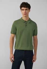 Zielona polo z krótkim rękawem o teksturowanym wykończeniu, z trzema guzikami i klasycznym kołnierzykiem. Noszona z ciemnoniebieskimi dżinsami.