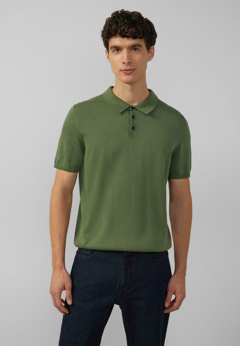 Zielona polo z krótkim rękawem o teksturowanym wykończeniu, z trzema guzikami i klasycznym kołnierzykiem. Noszona z ciemnoniebieskimi dżinsami.