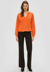 Oranje lange mouwen pullover met een V-hals en geplooide mouwen, gecombineerd met zwarte rechte broek en zwarte haklaarzen.