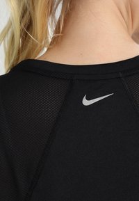 Camiseta deportiva negra con una textura de malla en la parte trasera, diseño de costuras planas y un pequeño logo plateado de Nike cerca del cuello.