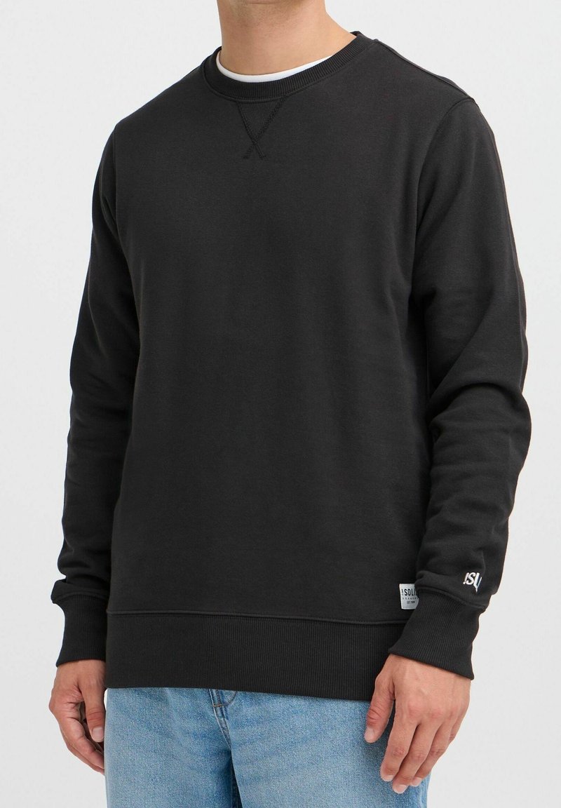 Homme portant un sweat-shirt noir à col rond avec poignets et ourlet côtelés, associé à un jean bleu clair, debout devant un fond uni.