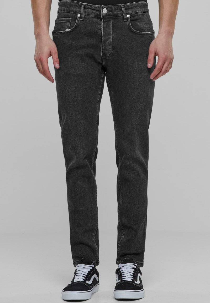 Fekete farmernadrág, slim fit szabással, öt zsebbel és ezüst színű részletekkel. Fekete és fehér sportcipőkkel viselve. Diszkrét kopás és sima felület jellemzi.