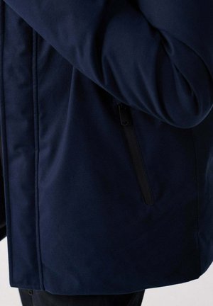 Veste bleu marine avec poche zippée sur le côté, montrée partiellement avec le bras plié sur la poitrine.