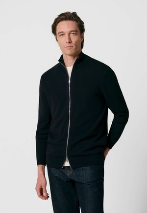 Uomo che indossa un maglione nero con zip e collo alto e jeans scuri, in piedi davanti a uno sfondo chiaro e uniforme.
