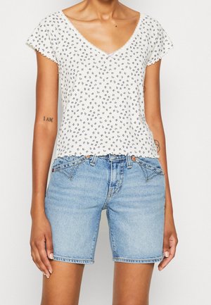Persoon draagt een wit kortärmelig bloementopje en lichtblauwe spijkershorts met voorzakken en knoopdetails, staand tegen een witte achtergrond.