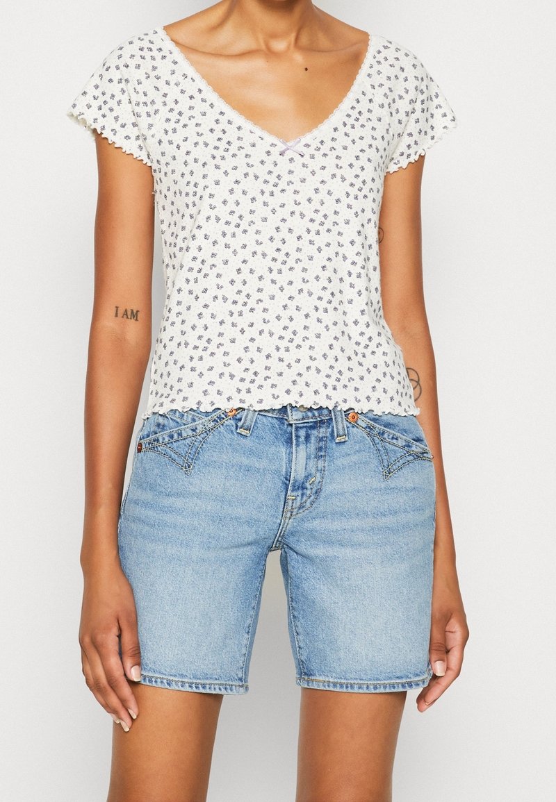 Personne portant un haut fleuri blanc à manches courtes et un short en jean bleu clair avec poches avant et détails de boutons, debout sur un fond blanc.