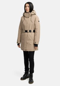 Parka matelassée beige avec capuche, ceinture à boucle noire, poches zippées et poignets élastiques. Portée par-dessus un pantalon slim noir et des bottes noires.