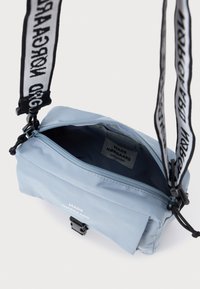 Jasnoniebieska torba na ramię, wykonana z miękkiego materiału. Posiada zamek błyskawiczny, regulowany pasek z logo oraz prostą metkę wewnętrzną.