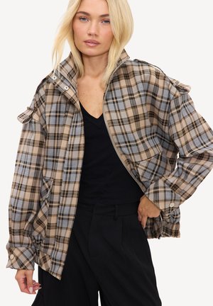 Femme aux cheveux blonds portant une veste zippée à carreaux beige et grise par-dessus un top noir et un pantalon noir taille haute.