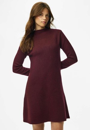 Young woman with long brown hair wearing a long-sleeve, knee-length, dark red knitted dress against a white background.