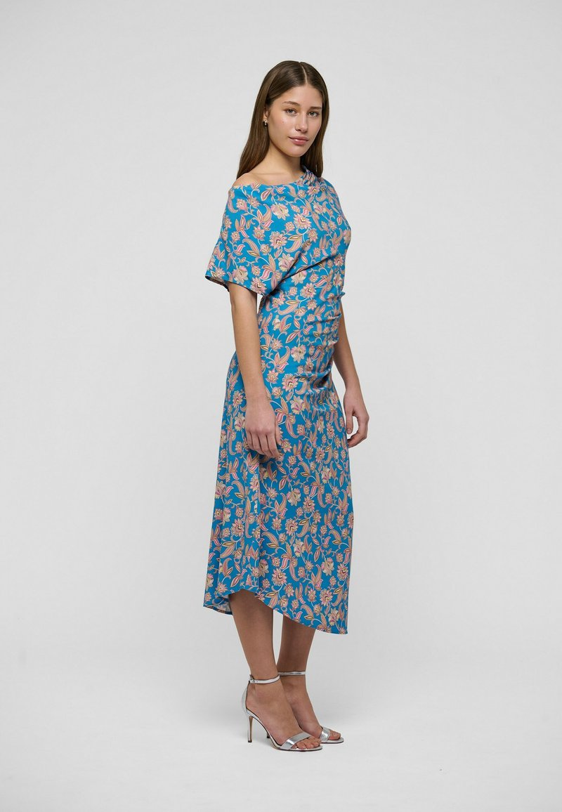 Femme debout portant une robe midi bleue à fleurs avec des manches courtes et des sandales à talons hauts argentées sur un fond uni.