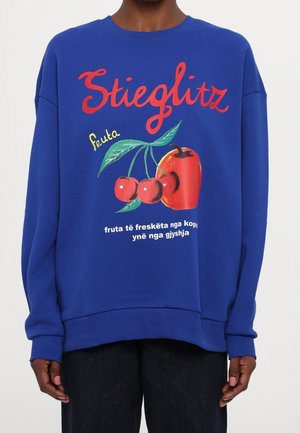 Niebieska bluza z czerwonym napisem "Stieglitz", ilustrowanymi wiśniami i czerwoną papryczką oraz białym albańskim tekstem o świeżych owocach.