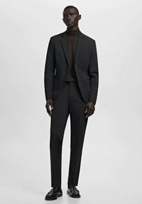 Lange man in een donker, op maat gemaakt pak met een bijpassende coltrui en zwarte loafers, staande tegen een effen lichte achtergrond.