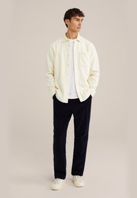Cream button-up shirt with a single chest pocket, paired with navy corduroy pants and white sneakers. Smooth fabric with relaxed fit.
