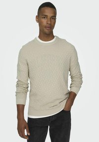 Pull en beige texturé à col rond présentant un motif en losanges et des poignets côtelés, superposé à un t-shirt blanc, associé à un jean noir.