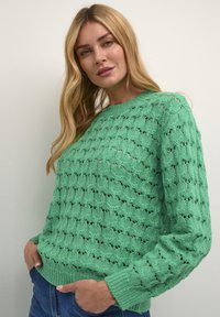Pulover tricotat verde deschis, cu un model din dantelă, mâneci lungi și guler și tiv cu nervuri. Asortat cu blugi lejeri albaștri.