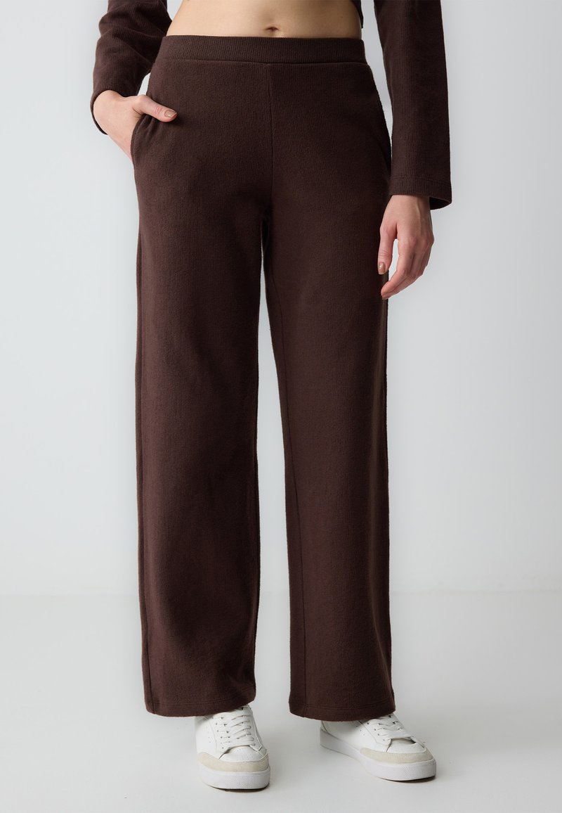 Pantaloni larghi marroni realizzati in tessuto testurizzato, con tasche laterali e una cintura a coste. Abbinati a sneakers bianche.