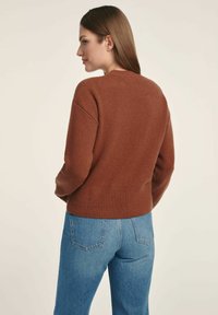 Brązowy sweter dzianinowy z prążkowanym wykończeniem na dole i rękawach, o luźnym kroju i okrągłym dekolcie. Połączony z jasnoniebieskimi dżinsami.