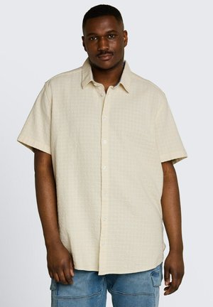Man draagt een beige korte mouwen button-up overhemd met textuurstof en blauwe cargoshorts, staand tegen een effen achtergrond.