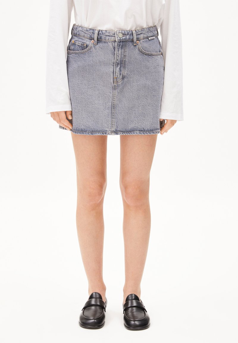 Jupe en denim avec un délavage bleu, design à cinq poches et coupe droite. Portée avec une chemise blanche à manches longues et des mocassins noirs.
