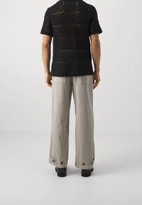 Camisa negra de punto de manga corta con tejido abierto y patrones cuadrados. Pantalones grises de pierna ancha con acentos de botón en los puños. Calzado negro.