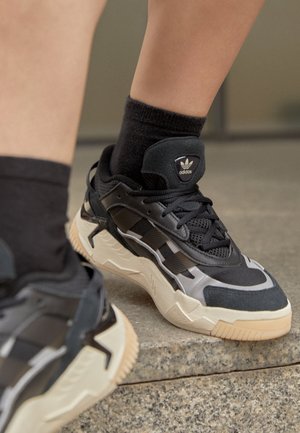 Pieds chaussés de baskets Adidas noires avec des semelles beige et des chaussettes noires, debout sur une marche en pierre à l'intérieur.