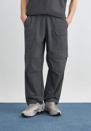 Person trägt dunkelgraue Cargo-Hose mit abnehmbaren Beinen und hellgraue Nike-Sneaker, steht auf einem blauen Teppich mit Händen in den Taschen.