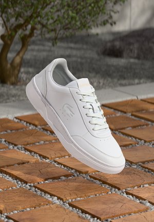 Baskets blanches en cuir avec un bout arrondi, un fermé à lacets, un branding subtil et une semelle en caoutchouc texturé, flottant au-dessus d'une surface en carreaux de bois.