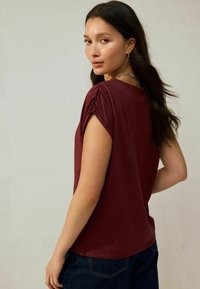 Burgundy short-sleeve top featuring gathered shoulder detail, soft fabric, relaxed fit, with a round neckline and curved hem.