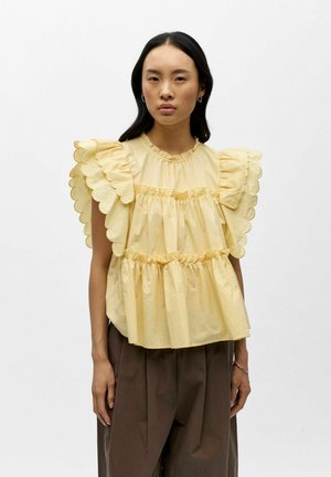 Femme portant un chemisier jaune sans manches avec des volants aux bords festonnés et un pantalon marron ample taille haute, debout devant un fond uni.