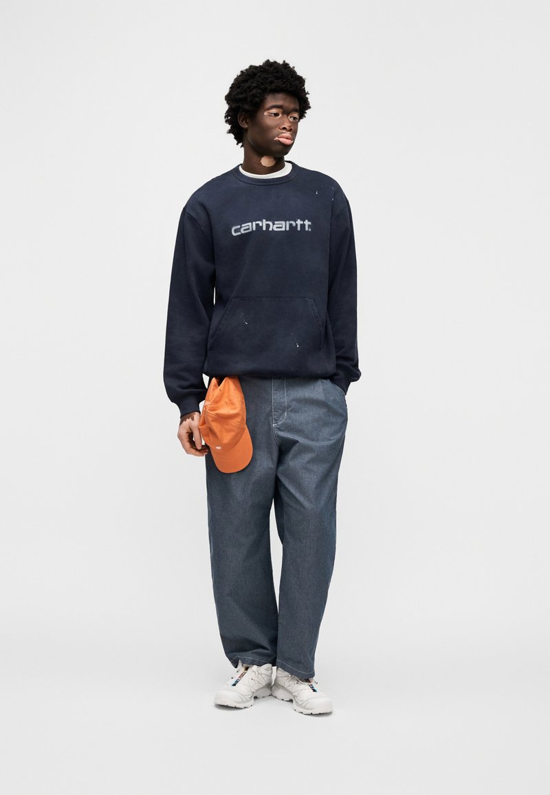 Marineblauer Sweatshirt mit "carhartt"-Logo, Jeanshose, orangefarbene Kappe und weiße Sneaker. Legerer Schnitt mit sichtbarer Textur und leichter Used-Optik.