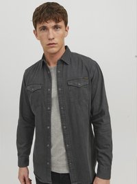Camisa de manga larga gris oscuro con cierre de botones y dos bolsillos en el pecho, llevada sobre una camiseta gris clara, con una textura suave y un ajuste casual.